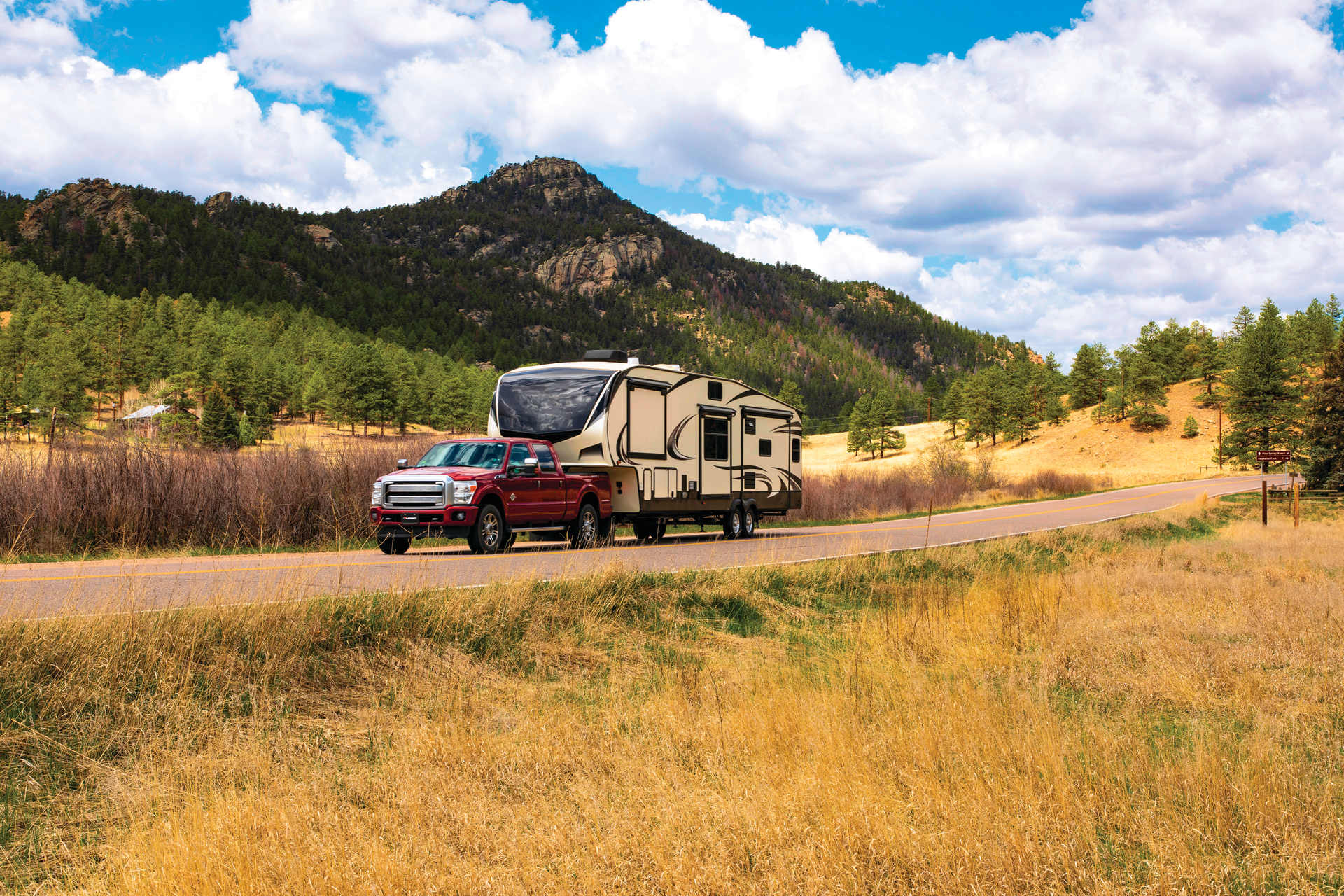 A red pickup truck towing a large beige camper trailer drives down a rural road with mountains and a partly cloudy sky in the background.